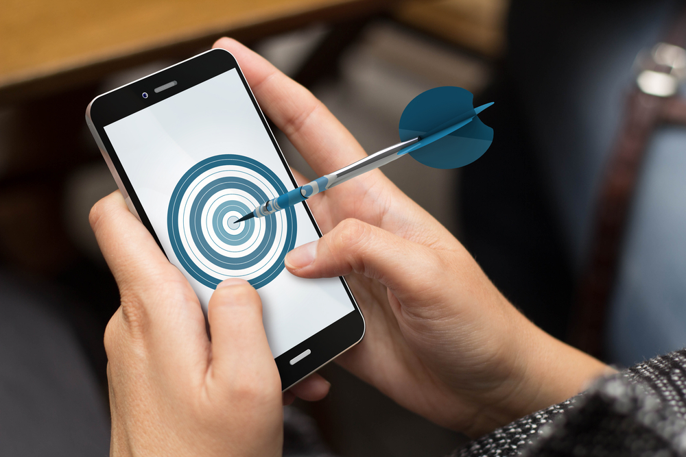 Person holds a cellphone in both hands, the screen displays a blue and white target, and a dart is stuck in the bulls-eye