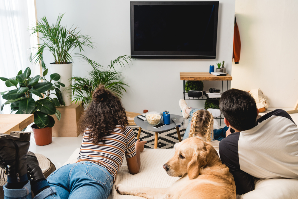 A group of people and a dog face toward a TV while laying on a couch with their backs to the camera