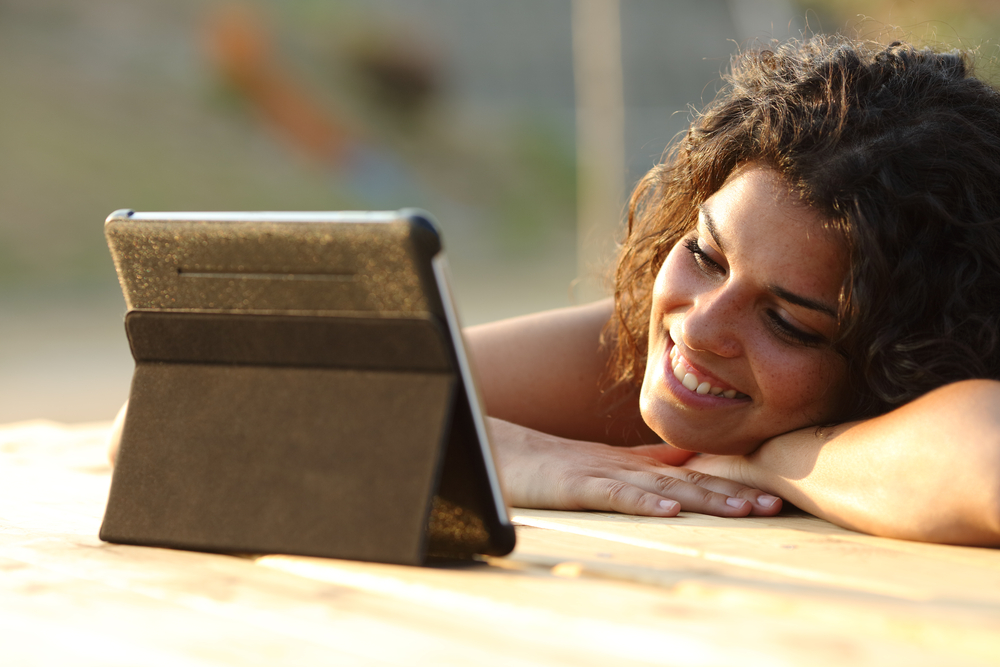 Woman leans on her hands at a table, looking at a propped up tablet